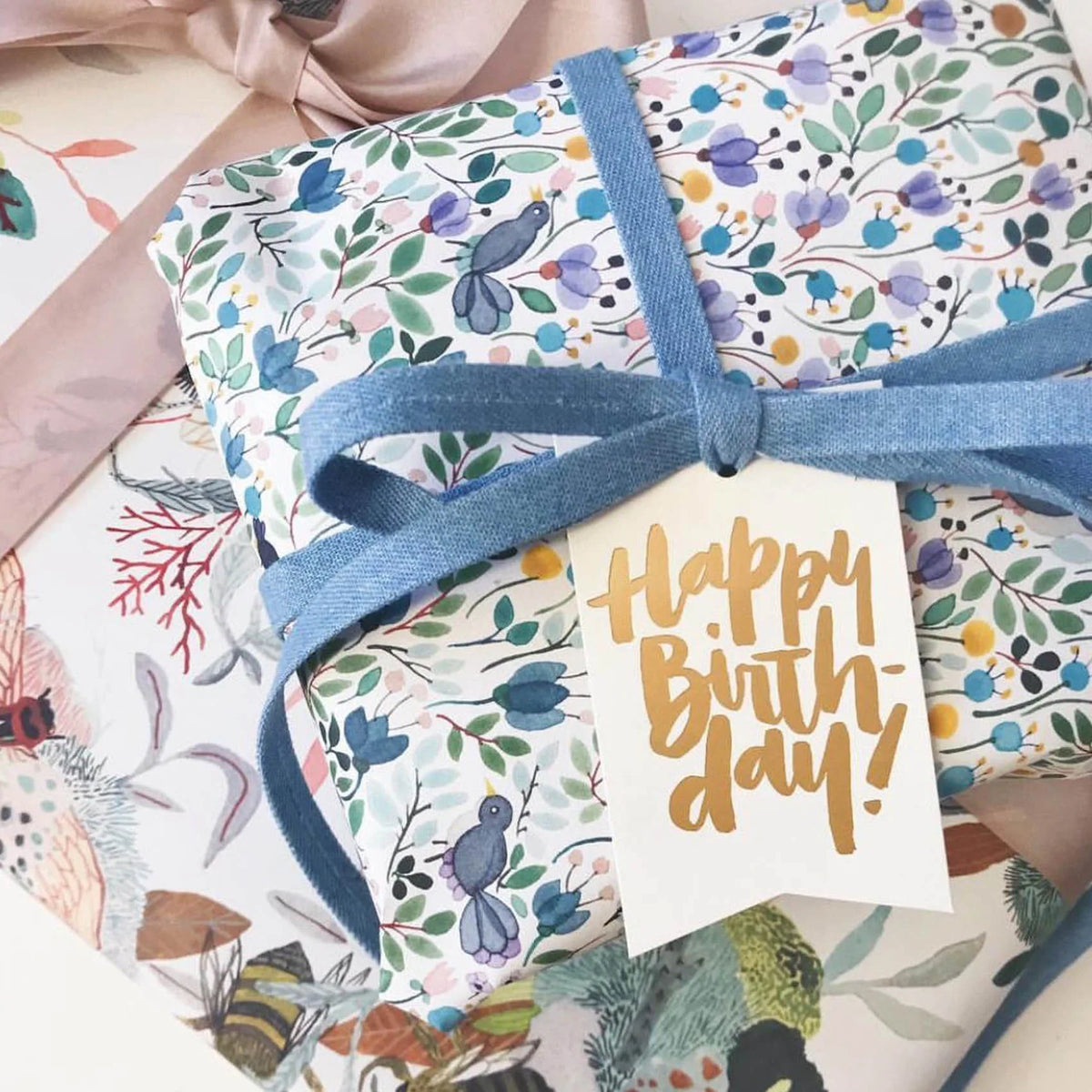 Cadeau d’anniversaire emballé dans un papier illustré de fleurs et d’oiseaux, avec ruban bleu et étiquette Happy Birthday