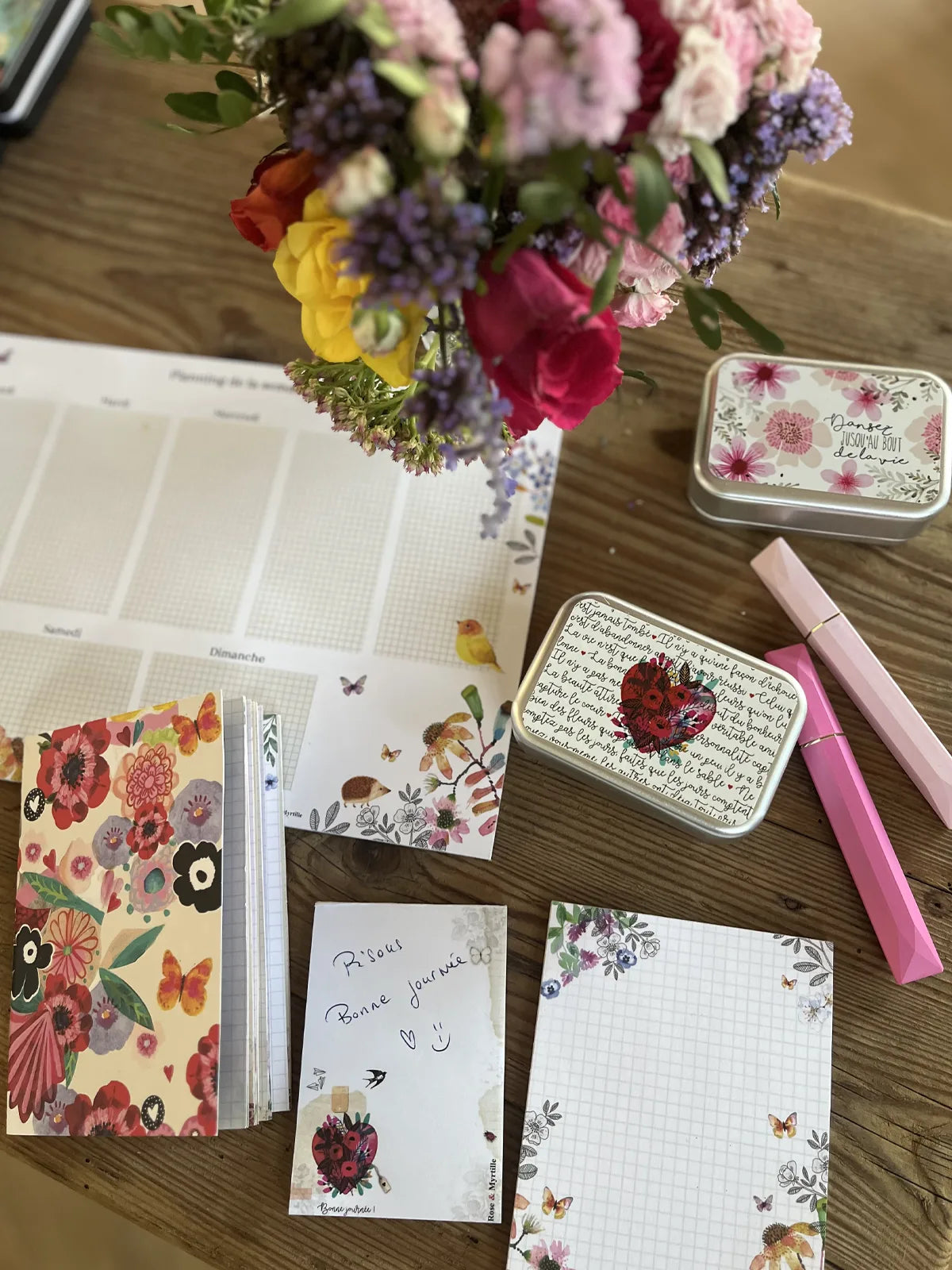 Créations illustrées Rose & Myrtille : planning, carnet fleuri, blocs-notes et boîtes métalliques disposés sur une table en bois